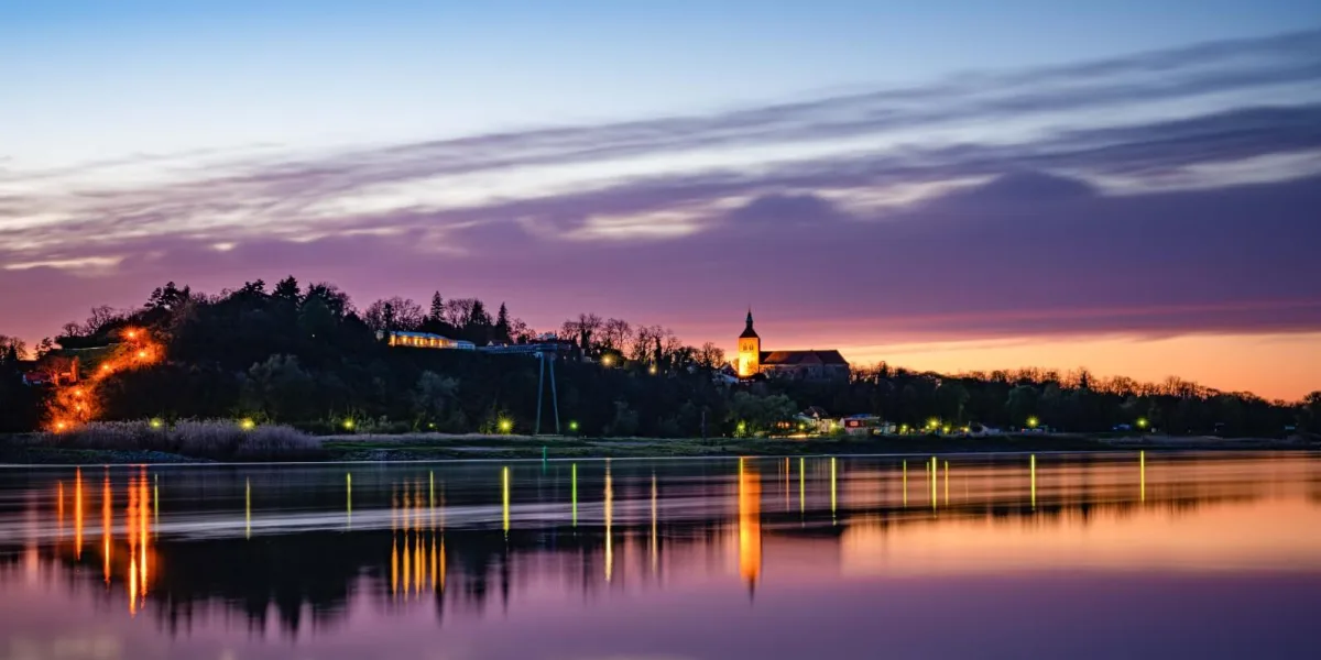 Blick über die Elbe auf Arneburg in der Abenddämmerung: Die beleuchtete Kirche Sankt Georg und das bewaldete Steilufer spiegeln sich im Wasser.